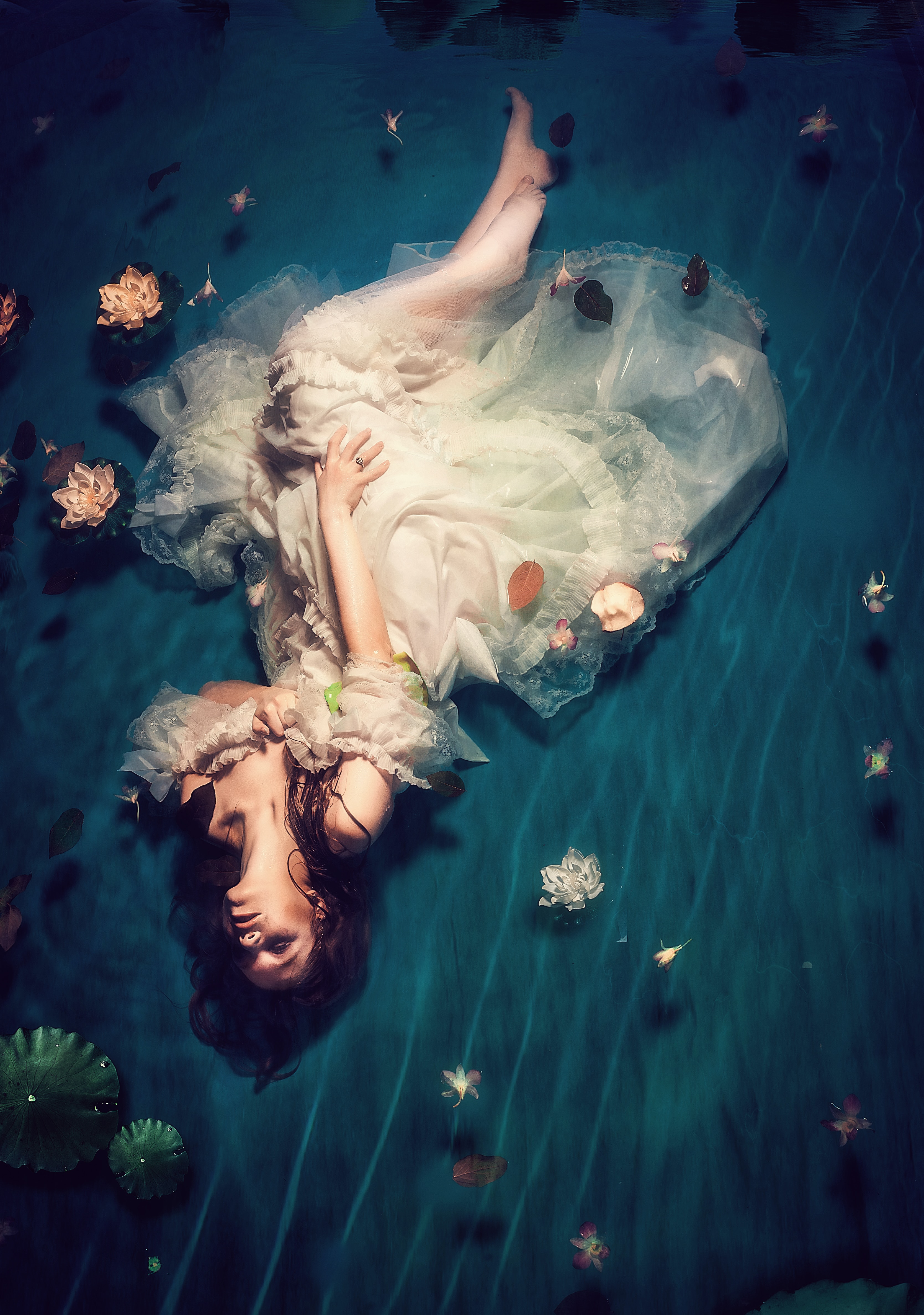 Woman in a white whimsical dress lying-in shallow water with flowers scattered around her.